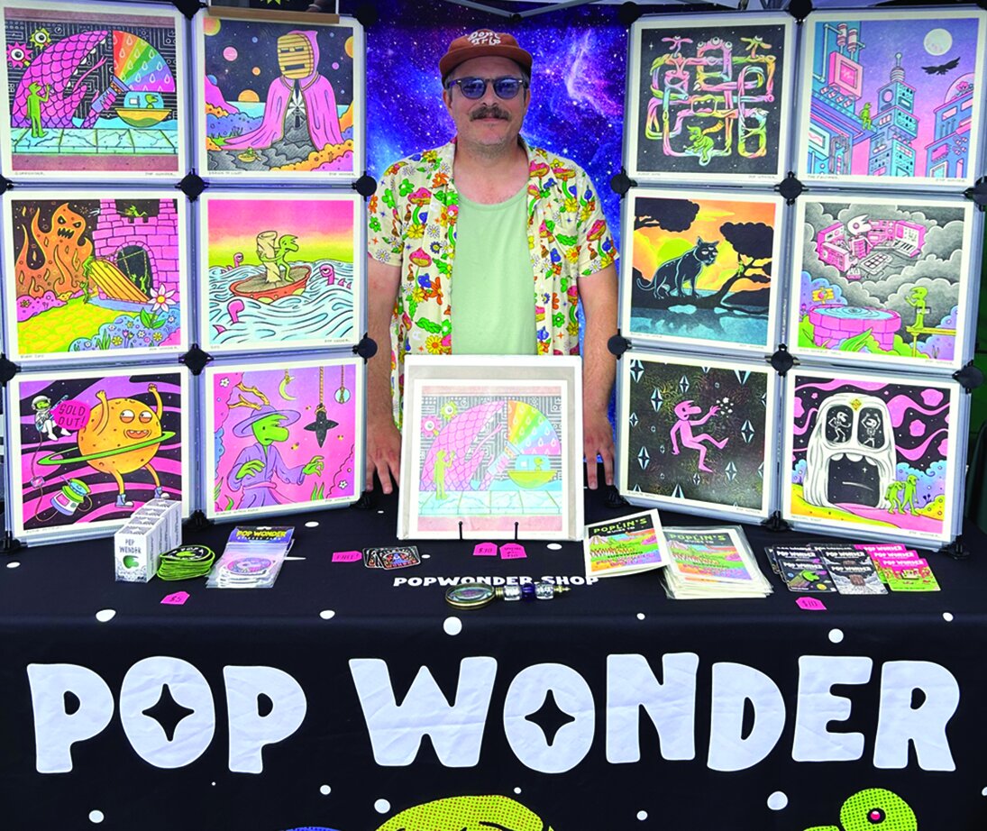 A vendor at the Pop Wonder booth surrounded by colorful art, mugs, and tote bags.