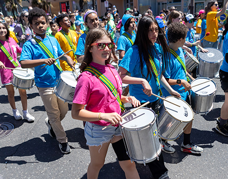 Children in pink and blue shirts marching with drums in a parade